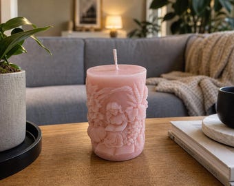 May include: A cylindrical light pink wax candle featuring intricate embossed relief carvings of a person reading a book surrounded by botanical elements, including wheat stalks and clusters of grapes, with a single centered cotton wick on top.