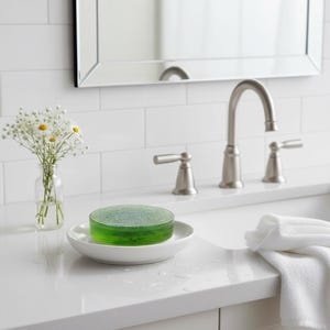 May include: A round, translucent green soap bar sits on a white ceramic soap dish. The soap dish is on a white countertop next to a white towel. A silver tap and a small vase of white flowers are also visible.