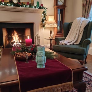 Könnte beinhalten: Gemütliches Wohnzimmer mit Kaminfeuer, grünem Sessel und Holztisch. Eine türkisfarbene Nussknackerfigur steht auf einem bordeauxfarbenen Tischläufer mit goldenen Fransen. Weihnachtsdekorationen schmücken den Kaminsims.