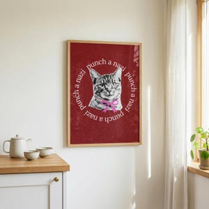May include: A framed wall art print featuring a dark red background with a central black and white illustration of a cat wearing a pink bow. The text "punch a nazi" is repeated in a circular pattern around the cat in white, sans-serif lettering.