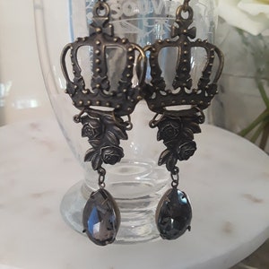 May include: A pair of antique-style earrings with intricate crown designs, rose details, and grey teardrop crystals. The earrings are made of bronze-coloured metal. The background is blurred. The earrings are displayed in front of a glass.
