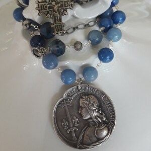 May include: A rosary with blue beads and a silver cross pendant. The rosary features a silver pendant with an image of Joan of Arc. The beads are a mix of light and dark blue, strung on a silver chain.