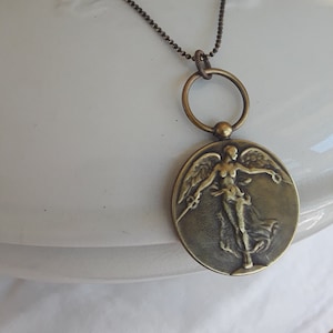 May include: Antique bronze-toned pendant necklace featuring a detailed relief of a winged figure. The pendant hangs from a delicate ball chain and a large ring. The necklace is displayed on a white ceramic surface.