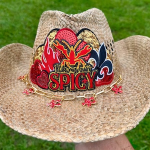 May include: A woven straw cowboy hat with a decorative patch. The patch features a red crawfish, flames, and the text "I like my bugs SPICY." A gold chain with red crawfish charms hangs from the brim. The hat is tan.