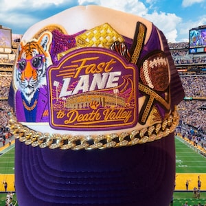 May include: A purple trucker hat featuring a large embroidered patch with the text Fast LANE to Death Valley, a tiger wearing sunglasses, a sequined football, and a gold-toned chain accent across the brim base with embedded clear rhinestones.