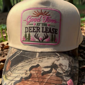 May include: A beige baseball cap with a camouflage brim and a rectangular patch featuring the text Good Times AT THE DEER LEASE in pink and brown. The patch includes embroidered graphics of a sun, pine trees, and deer antlers with pink hearts.