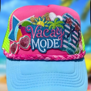 May include: A pink and light blue trucker hat featuring a braided pink trim and embroidered patches. The patches include a beach umbrella, heart-shaped sunglasses, a sun, a palm tree, and flip-flops, with the text Vacay MODE in pink and white.