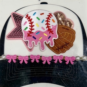 May include: A baseball cap with a white brim and black underside. The cap is decorated with a pink star, a baseball-shaped patch with sprinkles, and a brown baseball glove. A pink bow-shaped band is attached to the brim.