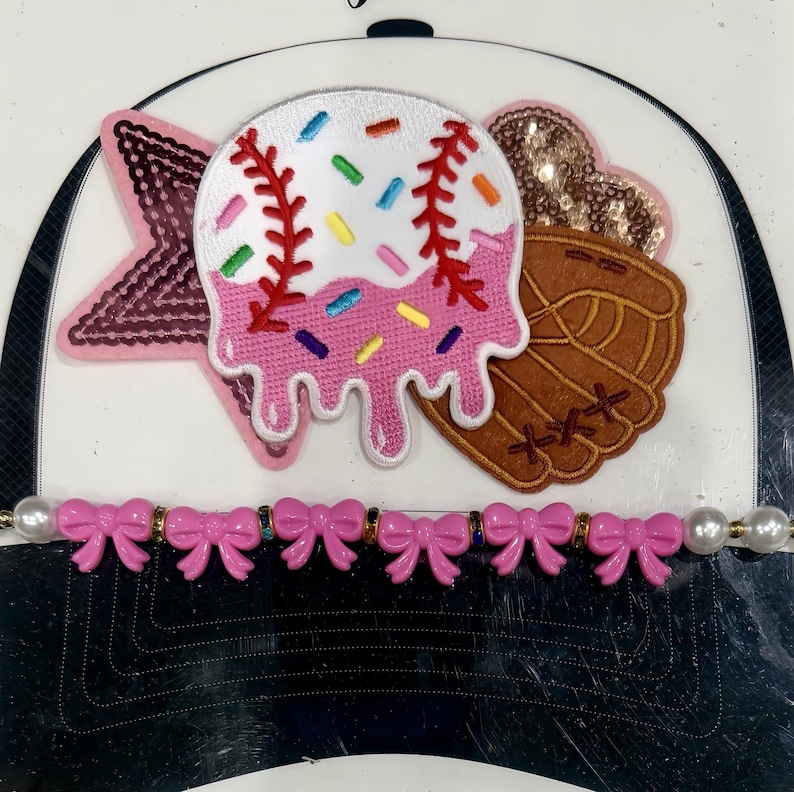 K&ouml;nnte beinhalten: Baseballkappe mit wei&szlig;em Schirm und schwarzer Unterseite. Die Kappe ist mit einem rosa Stern, einem baseballf&ouml;rmigen Aufn&auml;her mit Streuseln und einem braunen Baseballhandschuh verziert. Ein rosa Schleifenband ist am Schirm befestigt.