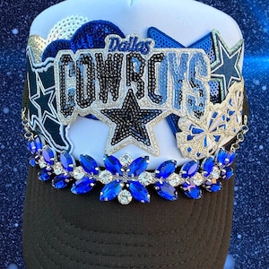 May include: A white and black trucker hat featuring a sequined Dallas Cowboys patch, blue and silver star appliqués, and a decorative chain across the brim adorned with blue and clear marquise-cut rhinestones in a floral pattern.