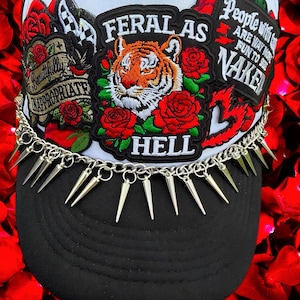 May include: A white and black trucker hat featuring multiple embroidered patches, including a tiger head with red roses and the text FERAL AS HELL, BEAUTIFULLY INAPPROPRIATE, and PEOPLE WITH TATTOOS ARE WAY MORE FUN TO SEE NAKED. A silver chain with spikes.