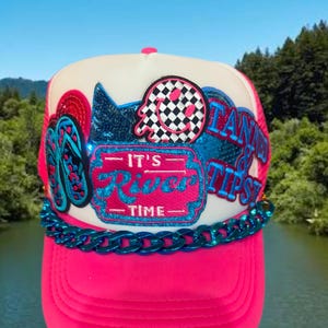 May include: A neon pink trucker hat featuring a white front panel with patches including a checkered smiley face, blue flip-flops, and a pink rectangular patch with the text IT'S RIVER TIME. A metallic blue chain trim accents the base of the crown.