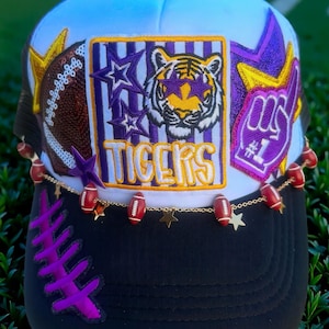 May include: A white and black trucker hat featuring a central patch with a tiger wearing star-shaped glasses, the text TIGERS, a football, and a number one foam finger. The hat is decorated with a gold chain featuring small football and star charms.