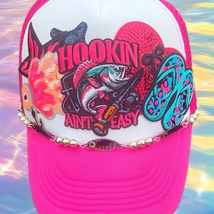 May include: A bright pink and white trucker hat featuring a central graphic of a fish and fishing rod with the text HOOKIN AINT EASY. The design includes sequined accents, a pair of blue and pink flip-flops, and a metallic beaded chain trim.