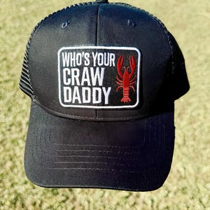 May include: Black trucker hat with a mesh back and a curved peak. The front has a rectangular patch with the text "WHO'S YOUR CRAW DADDY" and a red crawfish illustration. The hat is suitable for casual wear.