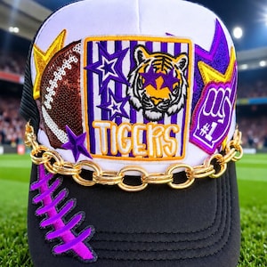 May include: A white and black baseball cap featuring a central patch with a tiger face, purple stars, and the text TIGERS. The cap includes a sequined football, a purple number one foam finger patch, a gold chain across the front, and purple laces.