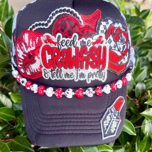 Op de afbeelding: Een zwarte truckerpet met een centrale geborduurde patch met de tekst "feed me CRAWFISH & tell me I'm pretty" in rood en zilver. De pet is versierd met paillettenlippen, een roos, een lippenstiftpatch en een kralenrand van rode en witte hartjes.