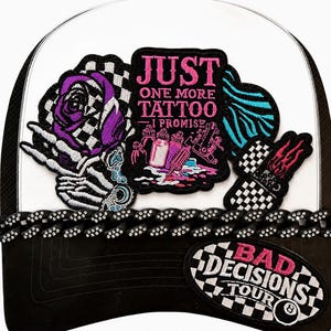 May include: A white and black baseball cap featuring embroidered patches including a skeleton hand holding a purple rose, a sign reading JUST ONE MORE TATTOO I PROMISE, and a patch with the text BAD DECISIONS TOUR and an eight ball graphic.