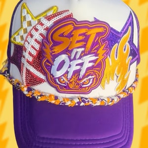 May include: A white and purple trucker hat featuring a central patch with the text SET IT OFF above a pair of eyes. The hat is decorated with a sequined football, a yellow star, a purple star, and a flame graphic, finished with a braided trim.