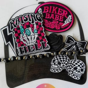 May include: A collection of patches and embellishments with a biker theme. Includes a patch with the text "RAISING HELL" and a skeleton hand, a patch that says "BIKER BABE", a black rose, a black bow, and two checkered skull charms.