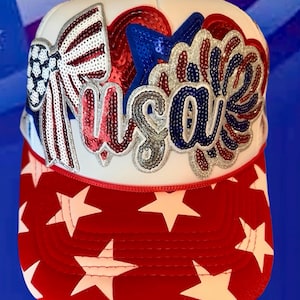 May include: A white trucker hat featuring a red brim with white stars. The front panel displays a large sequined applique with a bow, a blue star, and the text USA in red, white, and blue sequins, creating a patriotic and textured design.