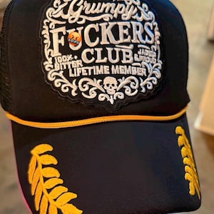 May include: Black trucker hat with a mesh back and a curved brim. The front patch reads "Grumpy F*ckers Club" in white script, with a skull and the words "Jaded & Proud" and "Lifetime Member". Gold leaf designs adorn the brim.
