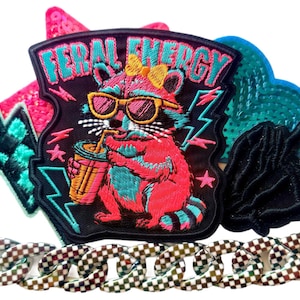 May include: An embroidered patch featuring a pink raccoon wearing yellow sunglasses and a yellow bow, drinking from a cup, with the text FERAL ENERGY. The patch is surrounded by other embroidered shapes including a teal lightning bolt and a black hand sign.