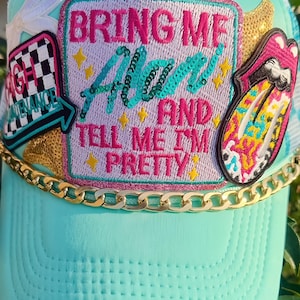 May include: A light turquoise trucker hat featuring a large central patch with the text BRING ME ALAN AND TELL ME I'M PRETTY in pink and teal sequins. The hat is decorated with a gold chain, a checkered patch, and a colorful lips patch.