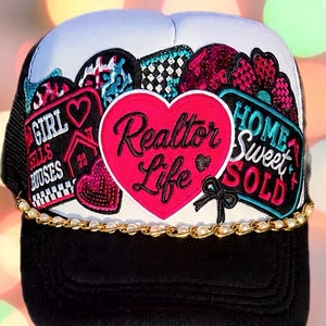 May include: A black and white trucker hat featuring multiple colorful patches, including a large pink heart with the text Realtor Life. Additional patches display the text Girl Sells Houses and Home Sweet Sold. A gold chain with white pearls accents the brim.
