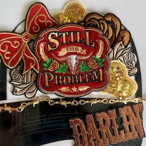 Op de afbeelding: Geborduurde patches met een Western-thema. Inclusief een rode patch met de woorden "STILL PROBLEM" en een schedel van een stier, een patch met het woord "DARLIN" en een gouden prikkeldraadontwerp. Andere patches zijn een rode strik en gouden paillettenharten.