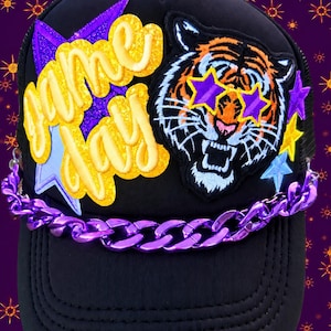 May include: A black trucker hat featuring a yellow glittery patch that reads Game Day layered over a purple star. Next to it is an embroidered tiger patch wearing purple star-shaped glasses. A metallic purple chain is attached across the front brim.