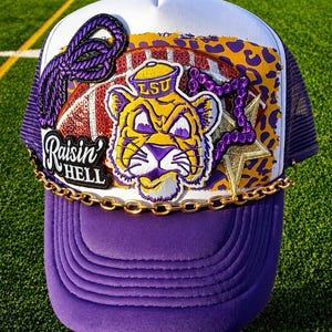 May include: A purple and white trucker hat featuring a central patch of a tiger mascot with the text LSU, a football graphic, a purple rope, a gold star, and a black patch with the text Raisin' HELL. A gold chain is draped across the front brim.