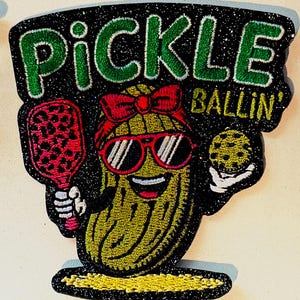 May include: A glittery patch featuring a cartoon pickle wearing sunglasses and a headband, holding a paddle and a pickleball. The text "PICKLE BALLIN'" is displayed above the pickle in green lettering.