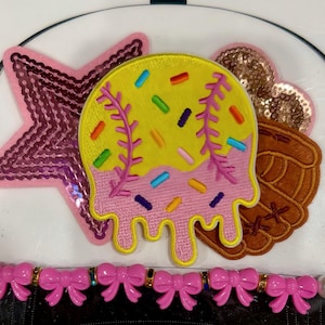 May include: A selection of decorative patches including a pink sequined star, a baseball-themed design with yellow, pink, and colourful sprinkles, a brown baseball glove, and a gold sequined heart. A pink bow and pearl trim is at the bottom.