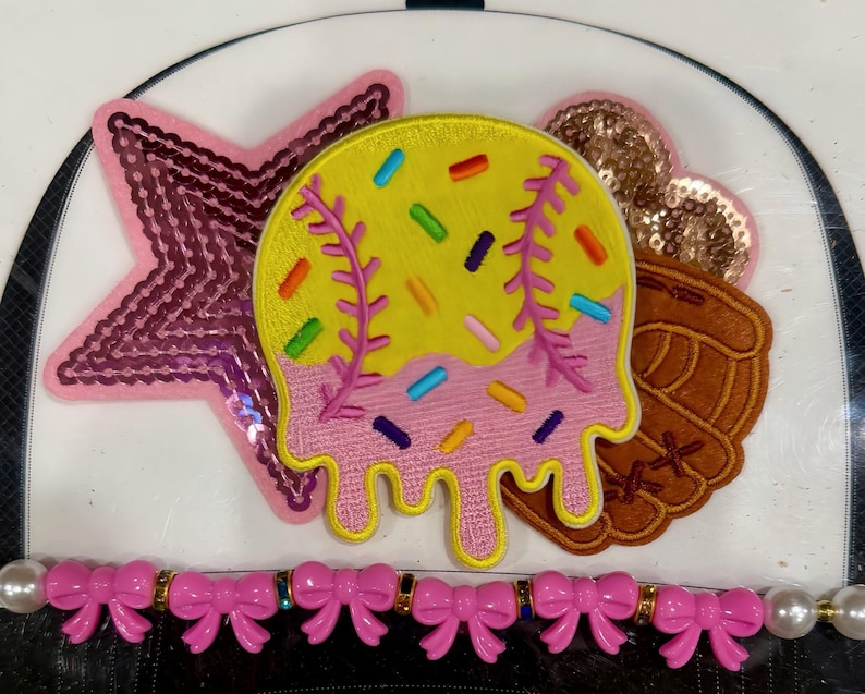 May include: A collection of decorative patches featuring a pink sequined star, a baseball-themed design with yellow, pink, and colorful sprinkles, a brown baseball glove, and a gold sequined heart. A pink bow and pearl trim is at the bottom.