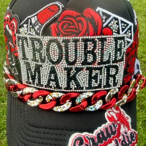 May include: Black trucker hat with the words "TROUBLE MAKER" in rhinestone lettering. The hat features red rose and playing card designs, a red chain, and a "Craw Baddie" patch.