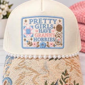 May include: A cream-colored baseball cap featuring a rectangular embroidered patch that reads PRETTY GIRLS HAVE GRANNY HOBBIES. The patch includes icons of a quilt block, yarn, and flowers. A string of white faux pearls lines the brim, which has a tan floral tapestry pattern.