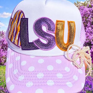 May include: A white trucker hat with a light purple brim featuring white polka dots. The front displays the letters LSU in purple and gold sequins, with a purple and gold striped heart patch. A strand of white faux pearls is attached to the side.