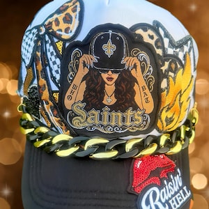 May include: A white and black trucker hat featuring a large central patch of a person wearing a cap with a fleur-de-lis, surrounded by leopard print, flames, and a red sequined lip patch with the text Raisin HELL. A thick gold and black chain.