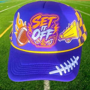 Puede incluir: Una gorra tipo trucker morada con un gráfico que incluye el texto SET IT OFF, un balón de fútbol americano marrón y un megáfono amarillo. La gorra tiene un cordón amarillo sobre la visera, un diseño de cordones blancos en el lateral y malla trasera.