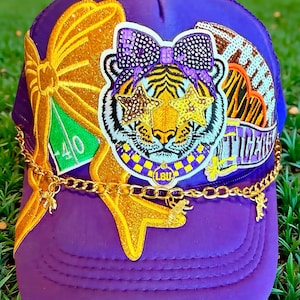 Può includere: Un cappellino trucker viola con una grande toppa centrale raffigurante una tigre con un fiocco di paillettes e occhiali a forma di stella. Il cappello è decorato con un fiocco glitterato dorato, una toppa da football e una catenella dorata.