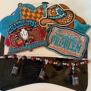 May include: A black hat brim adorned with embroidered patches featuring a cowboy hat, a skull, and text reading Chasin You Like a Shot of Whiskey and Somebody's Problem. A beaded strand with three miniature whiskey bottle charms is attached.