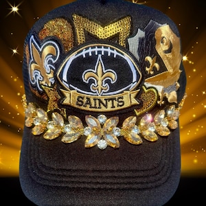 May include: A black baseball cap featuring a central patch with a football shape, a fleur-de-lis, and the text SAINTS. The cap is decorated with gold sequin stars, a fleur-de-lis patch, and a horizontal band of clear and amber-colored rhinestones.
