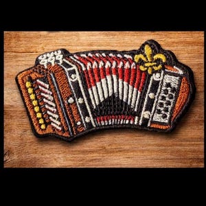 May include: Embroidered patch of an accordion in shades of brown, red, black, and white. The patch features a fleur-de-lis design and is set against a wooden background. The patch is approximately 4 inches wide.