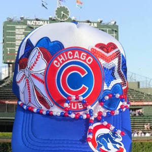 May include: A white and blue trucker hat featuring a circular Chicago Cubs logo patch, a large white bow with red stitching, a sequined star, and a blue heart. A strand of red, white, and blue beads drapes across the front, ending in a bear logo.