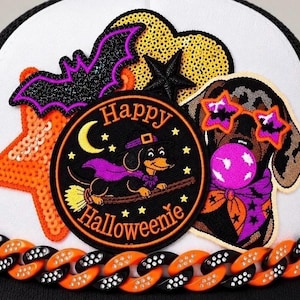May include: A white baseball cap featuring a circular patch with a dachshund on a broomstick and the text Happy Halloweenie, surrounded by sequined orange and yellow star patches, a glittery purple bat, and a dachshund wearing star glasses.