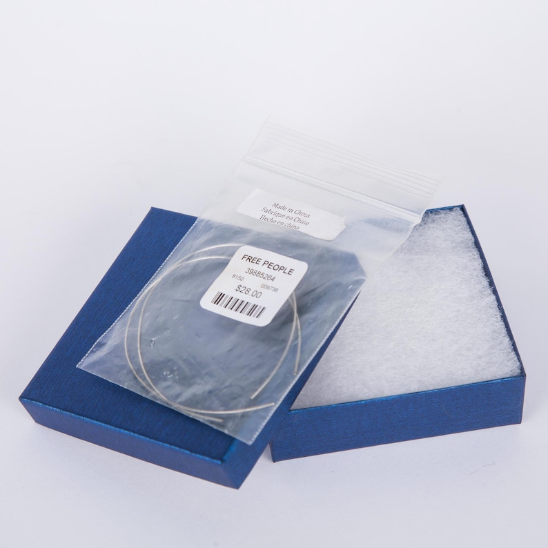 May include: Open blue jewelry box with a pair of thin, silver-colored bracelets inside a clear plastic bag. The bag has a label that reads "FREE PEOPLE" and a barcode. The box is lined with white padding.