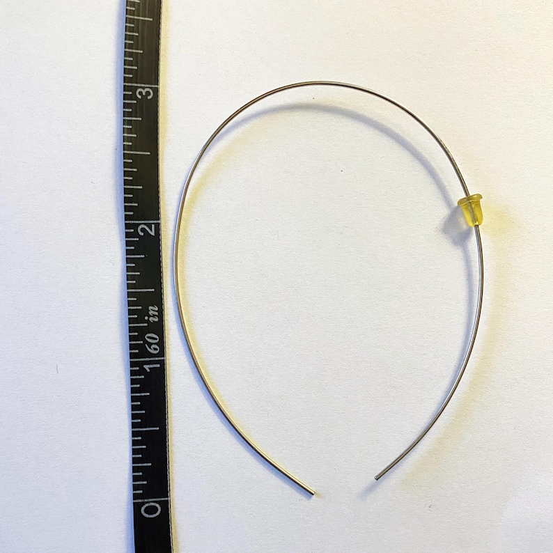 May include: A silver-colored wire earring with a yellow earring back. The earring is approximately 3 inches (7.6 cm) in diameter. The earring is on a white surface next to a black and white measuring tape.