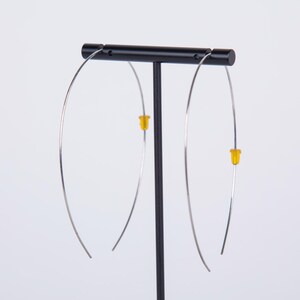 May include: A pair of silver-toned hoop earrings with yellow stoppers are displayed on a black stand. The earrings have a minimalist design, featuring a thin, curved wire that forms a large, open hoop. The yellow stoppers add a pop of color.