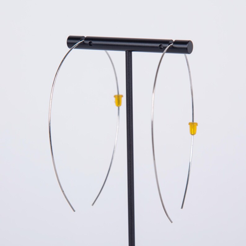 May include: A pair of silver-toned hoop earrings with yellow stoppers are displayed on a black stand. The earrings have a minimalist design, featuring a thin, curved wire that forms a large, open hoop. The yellow stoppers add a pop of color.